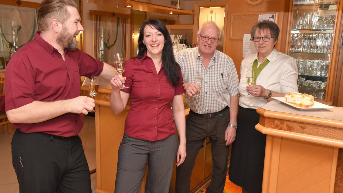Eine Gruppe von vier Personen steht an einer Bar und prostet mit Gläsern. Sie lächeln und wirken fröhlich in einer gemütlichen Umgebung. | © Teuschler-Mogg