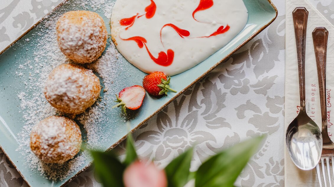 Ein Teller mit drei süßen Bällchen, bestreut mit Puderzucker, und einer cremigen Sauce mit rotem Muster. Nebenbei liegt eine frische Erdbeere. | © Teuschler_Mogg