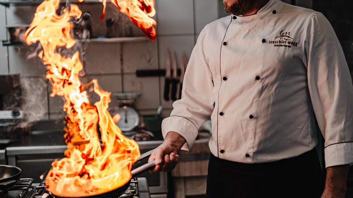 Ein Koch in einer modernen Küche, der eine Pfanne mit brennendem Essen schwenkt. Flammen steigen auf, während er konzentriert arbeitet. | © Teuschler_Mogg