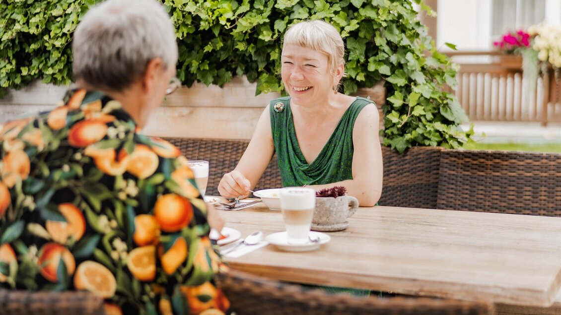 Zwei Frauen sitzen an einem Tisch im Freien und unterhalten sich. Im Hintergrund sind grüne Pflanzen und eine helle Fassade zu sehen. | © Teuschler_Mogg