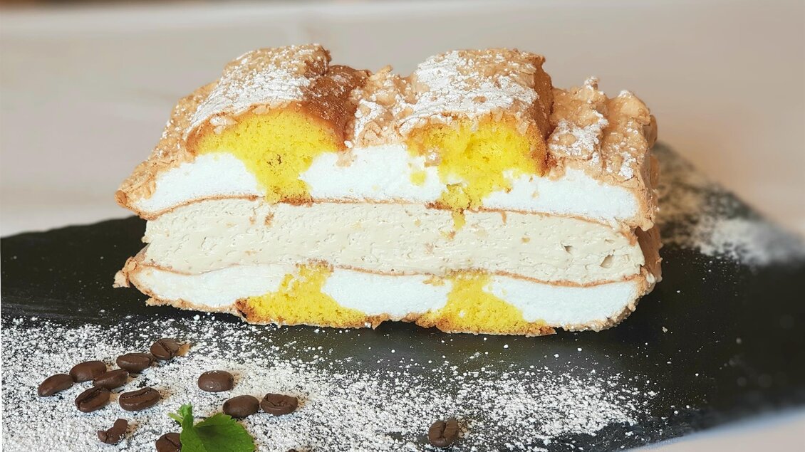 Ein köstlicher Biskuitkuchen mit Schichten auscremiger Füllung und fruchtiger Mango. Die Torte ist mit Puderzucker bestreut und auf einem schwarzen Teller angerichtet. | © Teuschler Mogg