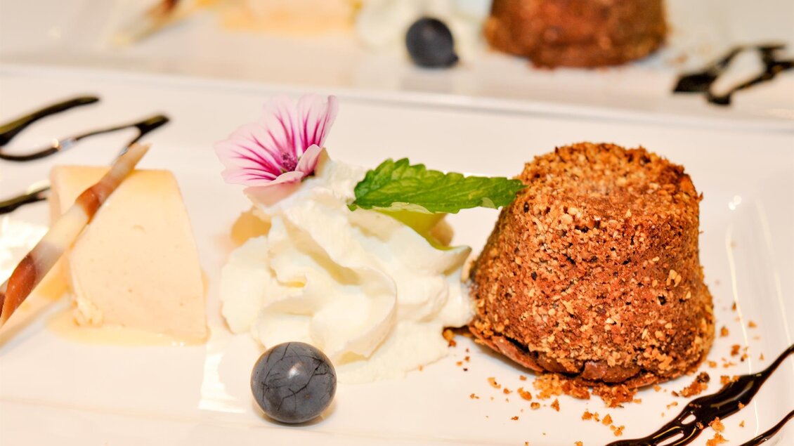 Ein verführendes Dessert auf einem weißen Teller. Es besteht aus einem Schokoladengebäck, einer Kugel Eis, Sahne, einer Blaubeere und einem essbaren Blumenzweig. | © Teuschler Mogg