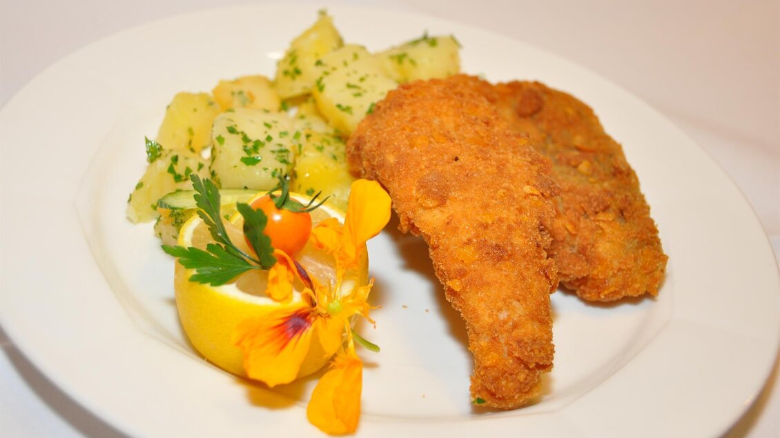 Ein Teller mit knusprig gebratenem Fischfilet, serviert mit Kartoffelsalat und einer Zitronenscheibe. Dazu eine essbare Blüte zur Dekoration. | © Teuschler Mogg