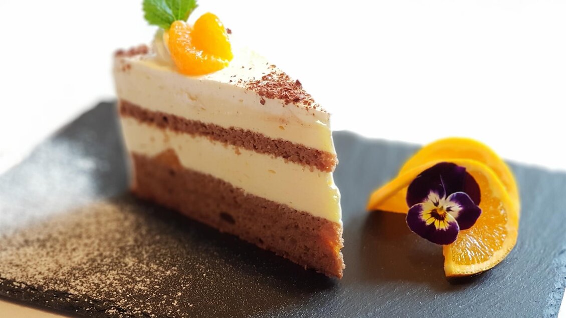 Ein Stück Schokoladenkuchen mit einer cremigen Schicht und dekoriert mit einer Orange und einer essbaren Blume. Der Kuchen liegt auf einem schwarzen Teller. | © Teuschler Mogg