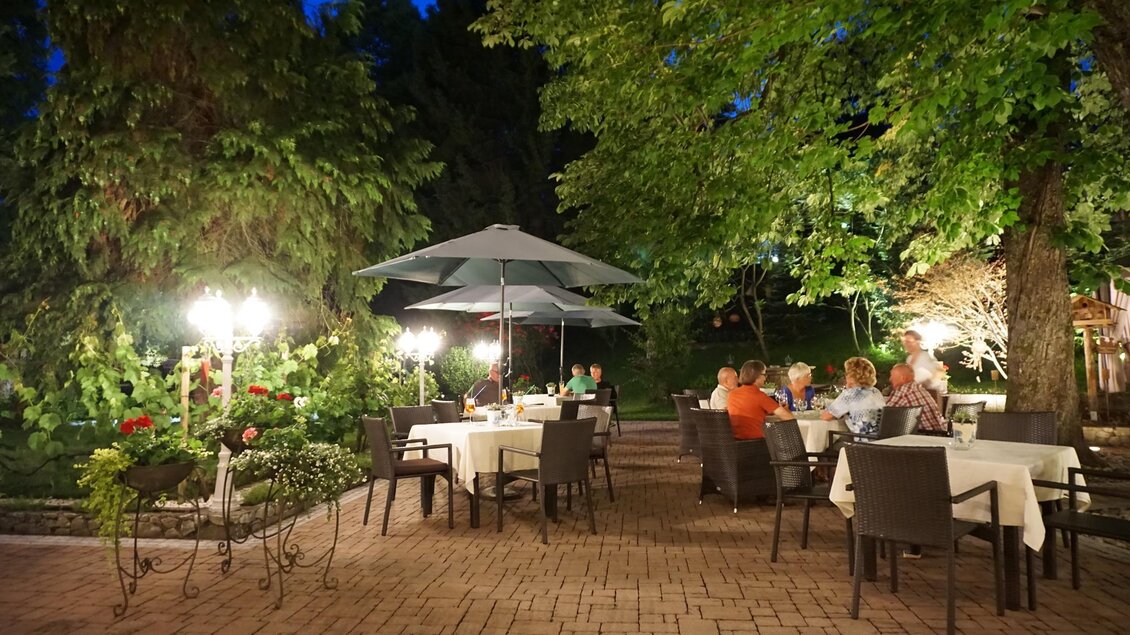 Ein gemütliches Restaurant im Freien mit Tischen und Stühlen unter großen Bäumen. Es ist Abend, und die Atmosphäre wird von sanften Lichtern und einer entspannten Stimmung geprägt. | © Kloepferkeller