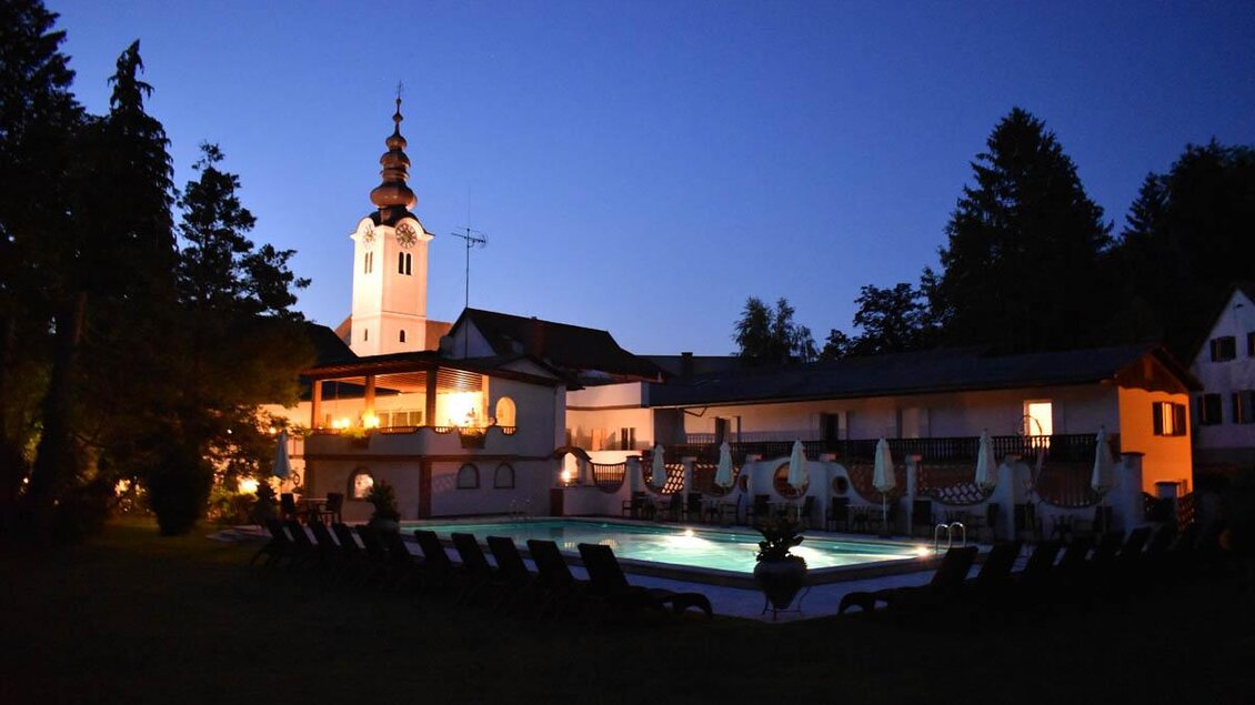 Ein Hotel mit beleuchtetem Pool und einer Kirche im Hintergrund bei Nacht. Der Garten ist ruhig und einladend. | © Desiree Zwanzleitner