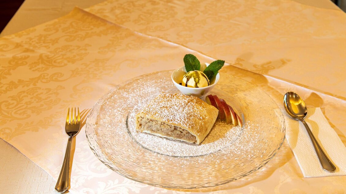 Ein Stück Apfelstrudel mit Puderzucker und einer kleinen Portion Eiscreme auf einem eleganten Teller. Der Tisch ist mit einer feinen Tischdecke ausgestattet. | © Hotel Neuhold