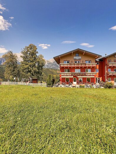 Ein rotes Gebäude mit mehreren Etagen steht vor einer grünen Wiese. Im Hintergrund sind Berge und ein blauer Himmel mit einigen Wolken zu sehen. | © Algo