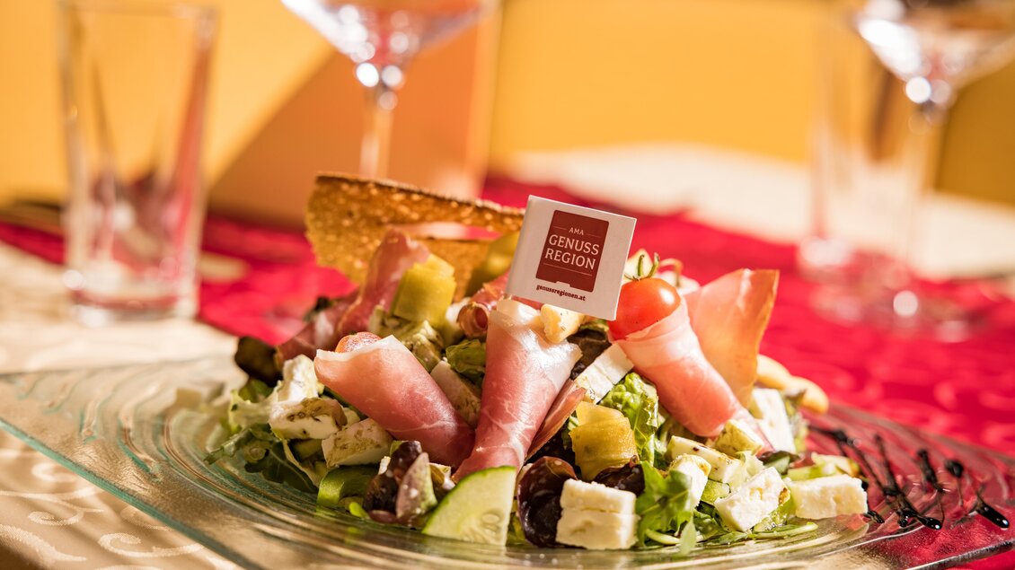Ein frischer Salat mit Schinken, Käse und buntem Gemüse. Der Teller ist elegant angerichtet und hat ein kleines Schild mit der Aufschrift "GENUSS REGION". | © AMA