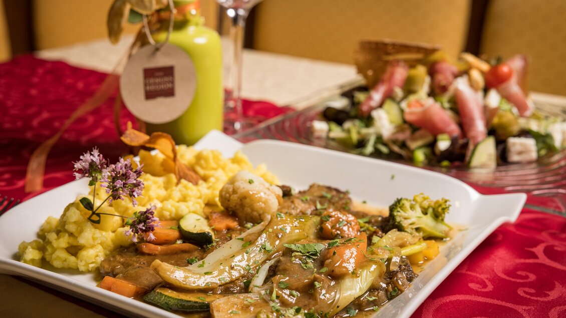 Ein elegantes Gericht mit Gemüse und Fleisch auf einem weißen Teller, garniert mit frischen Kräutern. Im Hintergrund befindet sich ein bunter Salat und ein Getränk. | © AMA