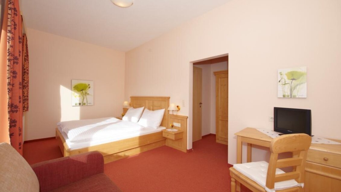 Ein helles Zimmer mit einem Doppelbett, einem Schreibtisch und einem Sessel. Die Wände sind in sanften Farben gehalten, und es gibt dekorative Bilder an der Wand. | © Algo