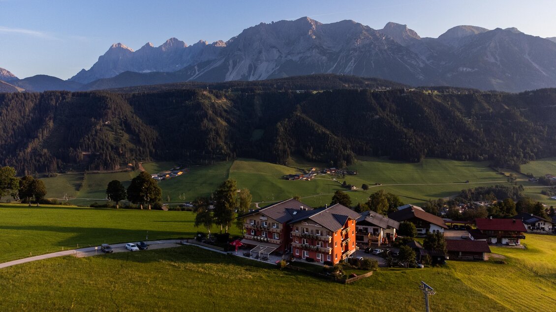 Eine malerische Landschaft mit sanften Hügeln und majestätischen Bergen im Hintergrund. Im Vordergrund steht ein charmantes Gebäude inmitten von grünen Wiesen. | © Julius Kotus