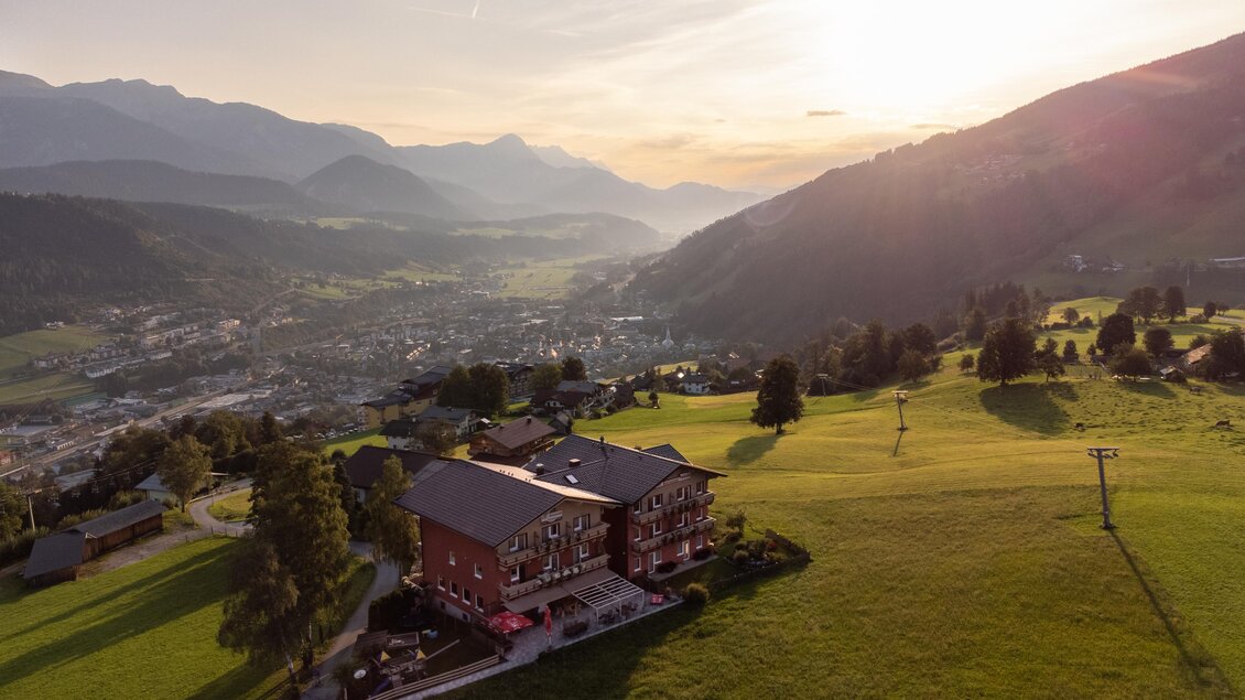 Eine malerische Landschaft mit einem Gebäude in der Mitte, umgeben von grünen Wiesen und Bergen im Hintergrund. Die Sonne geht unter und taucht die Szene in warmes Licht. | © Julius Kotus