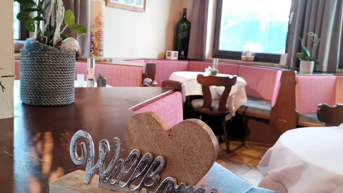 Eine einladende Restaurantatmosphäre mit einem willkommenen Schild aus Holz. Am Tisch stehen Pflanzen und das Licht sorgt für Gemütlichkeit. | © Hotel Pariente