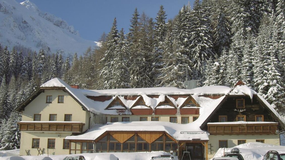 Ein schneebedecktes Hotel umgeben von hohen Tannen und Bergen. Die Parkplätze sind ebenfalls mit Schnee bedeckt. | © Präbichlerhof