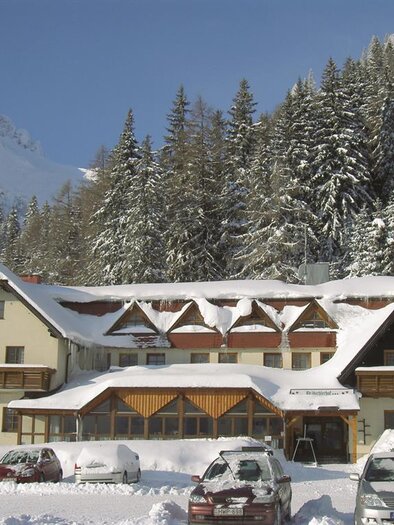 Ein schneebedecktes Hotel umgeben von hohen Tannen und Bergen. Die Parkplätze sind ebenfalls mit Schnee bedeckt. | © Präbichlerhof