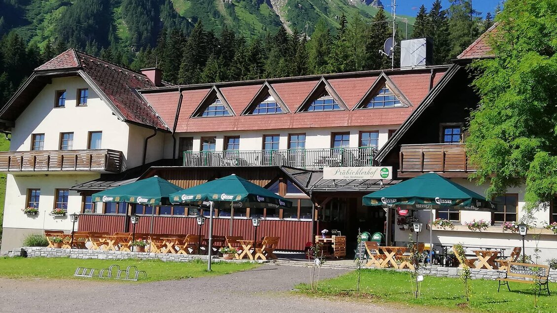 Ein gemütliches Gasthaus in den Bergen mit einer einladenden Terrasse. Im Hintergrund sind grüne Wälder und majestätische Berge zu sehen. | © Strohmaier