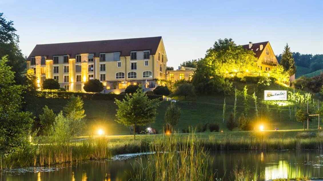 Ein ruhiges Hotelgebäude liegt am Ufer eines Teichs, umgeben von Bäumen und grüner Landschaft. Die Abendbeleuchtung verleiht der Szene eine einladende Atmosphäre. | © Hotel-Restaurant Staribacher