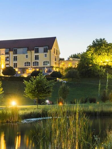 A quiet hotel building is located on the edge of a pond, surrounded by trees and green landscape. The evening lighting gives the scene an inviting atmosphere. | © Hotel-Restaurant Staribacher