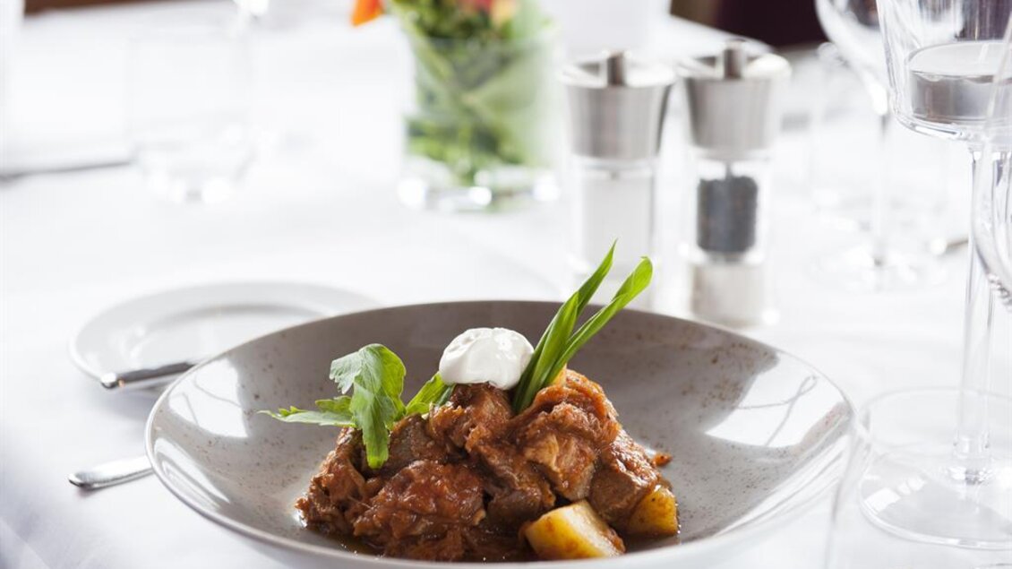 Ein köstliches Gericht in einer eleganten Schüssel, garniert mit frischen Kräutern und einem Klecks Sahne. Der Tisch ist schön gedeckt mit Gläsern und Blumendekoration. | © Hotel-Restaurant Staribacher