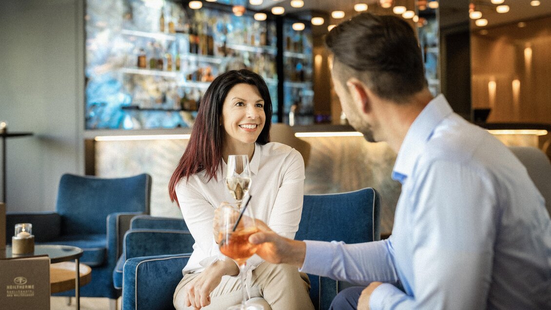 Zwei Personen sitzen in einer eleganten Lounge und unterhalten sich. Eine Frau lächelt und hält ein Getränk in der Hand. | © Heilthermen Resort BW