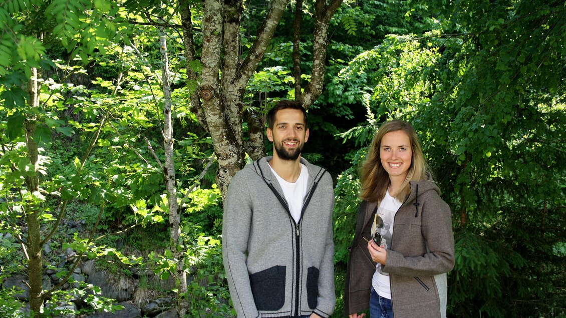 Ein Paar steht lächelnd in einem grünen Wald. Im Hintergrund sind Bäume und eine natürliche Umgebung zu sehen. | © Huber Strick- und Walkwaren