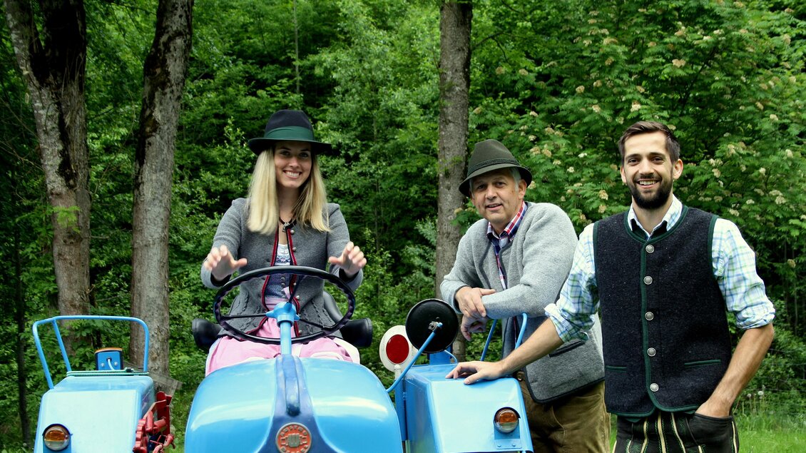 Drei Personen stehen neben einem blauen Traktor in einer grünen Landschaft. Die Männer tragen traditionelle Kleidung, während die Frau einen Hut und einen Pulli trägt. | © Huber Strick- und Walkwaren