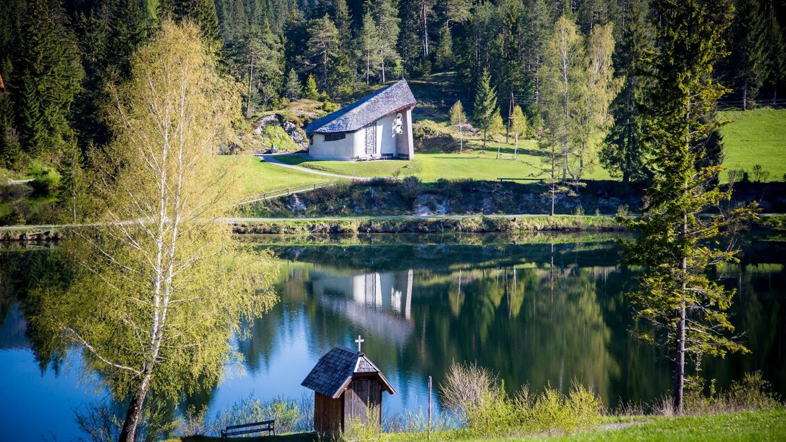 Eine malerische Landschaft mit einem ruhigen See und grünen Wiesen. Im Hintergrund steht ein modernes Haus und kleine Bäume spiegeln sich im Wasser. | © TV Hochsteiermark/Fred Lindmoser