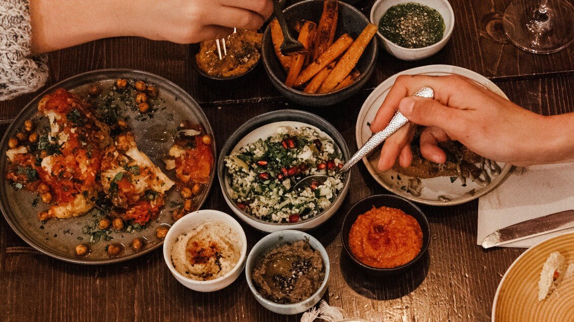Ein gedeckter Tisch mit verschiedenen köstlichen Gerichten und Beilagen. Hände, die sich dem Essen nähern, schaffen eine gesellige Stimmung. | © Hummel Graz