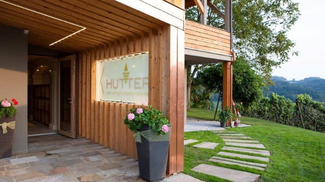 Ein modernes Holzgebäude mit einer einladenden Eingangstür und einem gepflegten Garten. Blumen in Töpfen und ein Steinweg ergänzen die gemütliche Atmosphäre. | © Weingut Hutter