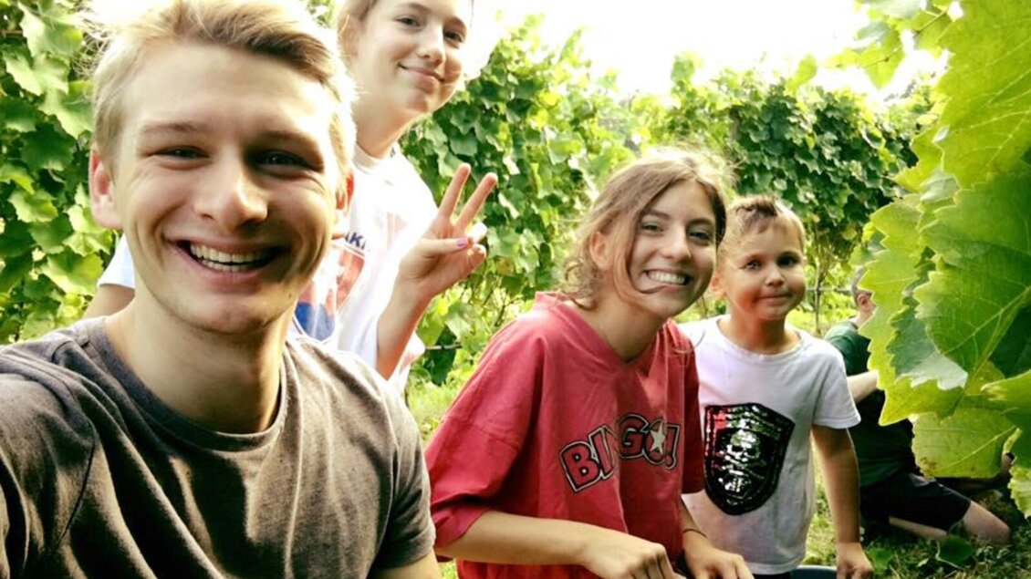 Eine Gruppe junger Menschen lächelt fröhlich in einem Weinberg. Im Hintergrund sind grüne Weinreben zu sehen. | © Weingut Hutter