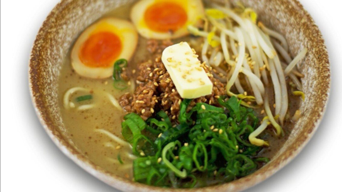 Eine Schüssel Ramen mit Brühe, frischem Gemüse, Sesam und zwei halben weichgekochten Eiern. Ein Stück Butter und grüne Zwiebeln sind als Garnitur hinzugefügt. | © Ichi-Go Ichi-E