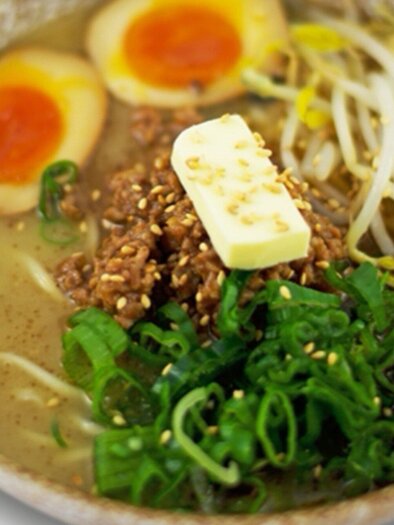 A bowl of ramen with broth, fresh vegetables, sesame, and two half-soft-boiled eggs. A piece of butter and green onions are added as garnish. | © Ichi-Go Ichi-E
