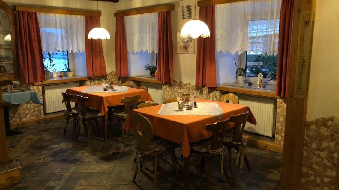 Ein gemütliches Restaurant mit Holzbalkendecke und Fenstern mit Vorhängen. Die Tische sind mit orangefarbenen Tischdecken gedeckt und es gibt warmes Licht von den Lampen.