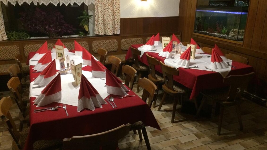 Ein gemütlicher Gastraum mit mehreren Tischen, die mit roten Tischdecken und gefalteten Servietten dekoriert sind. Im Hintergrund gibt es ein großes Fenster und ein Fischbecken.