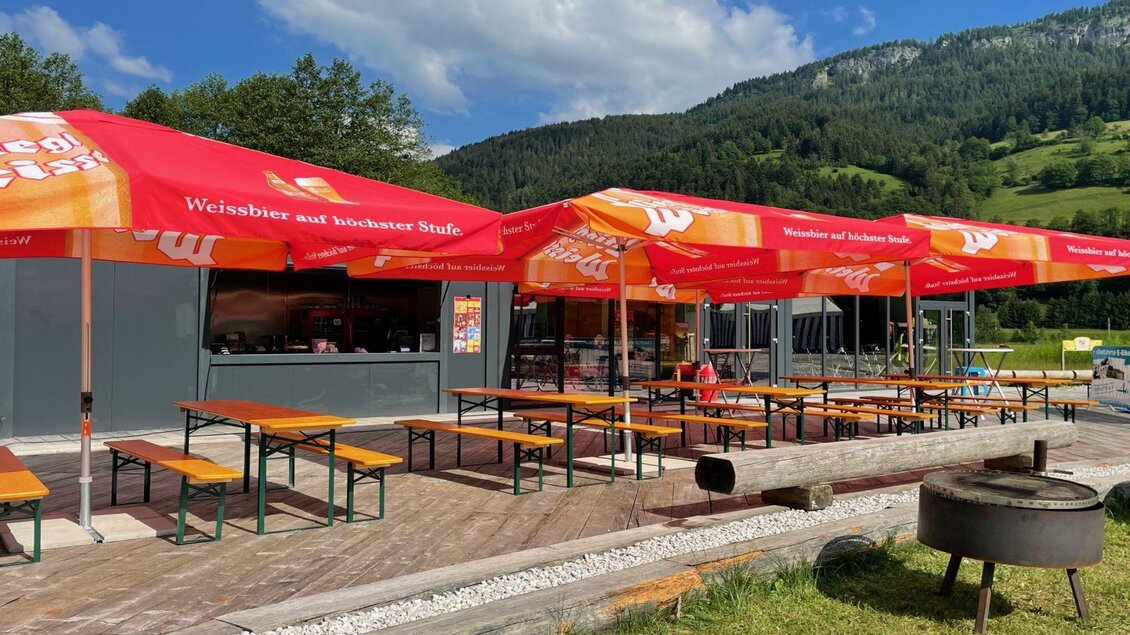 Ein gemütlicher Außenbereich mit langen Bänken und Tischen unter roten Sonnenschirmen. Im Hintergrund sind grüne Hügel und blauer Himmel zu sehen. | © Auszeit Ausseerland