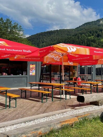 Ein gemütlicher Außenbereich mit langen Bänken und Tischen unter roten Sonnenschirmen. Im Hintergrund sind grüne Hügel und blauer Himmel zu sehen. | © Auszeit Ausseerland