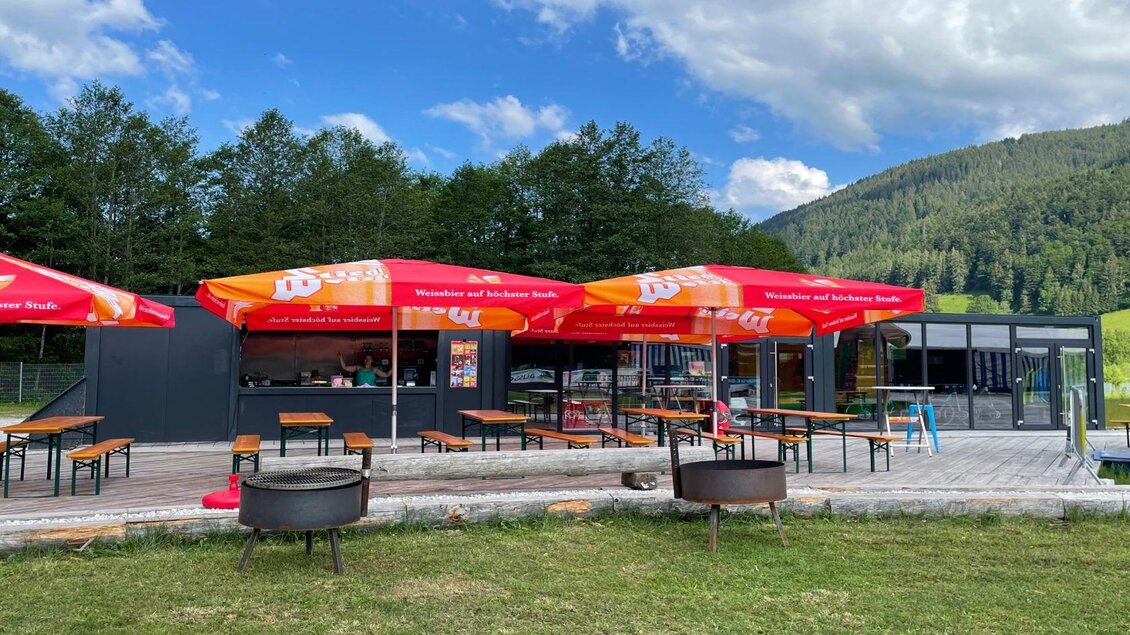 Ein gemütlicher Außenbereich mit Holz-Bänken und roten Sonnenschirmen. Im Hintergrund sind Bäume und Berge zu sehen, unter einem klaren Himmel. | © Auszeit Ausseerland