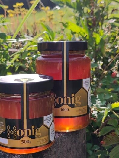 Zwei Honiggläser stehen auf einem Holzblock inmitten einer blühenden Gartenlandschaft. Die Gläser sind orange und goldfarben, umgeben von grünen Pflanzen. | © Miriam Wachter