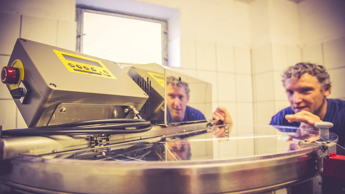 Ein Mann beobachtet gespannt eine Maschine in einem hellen Raum. Die Maschine hat eine glänzende Oberfläche und ist Teil eines Produktionsprozesses.