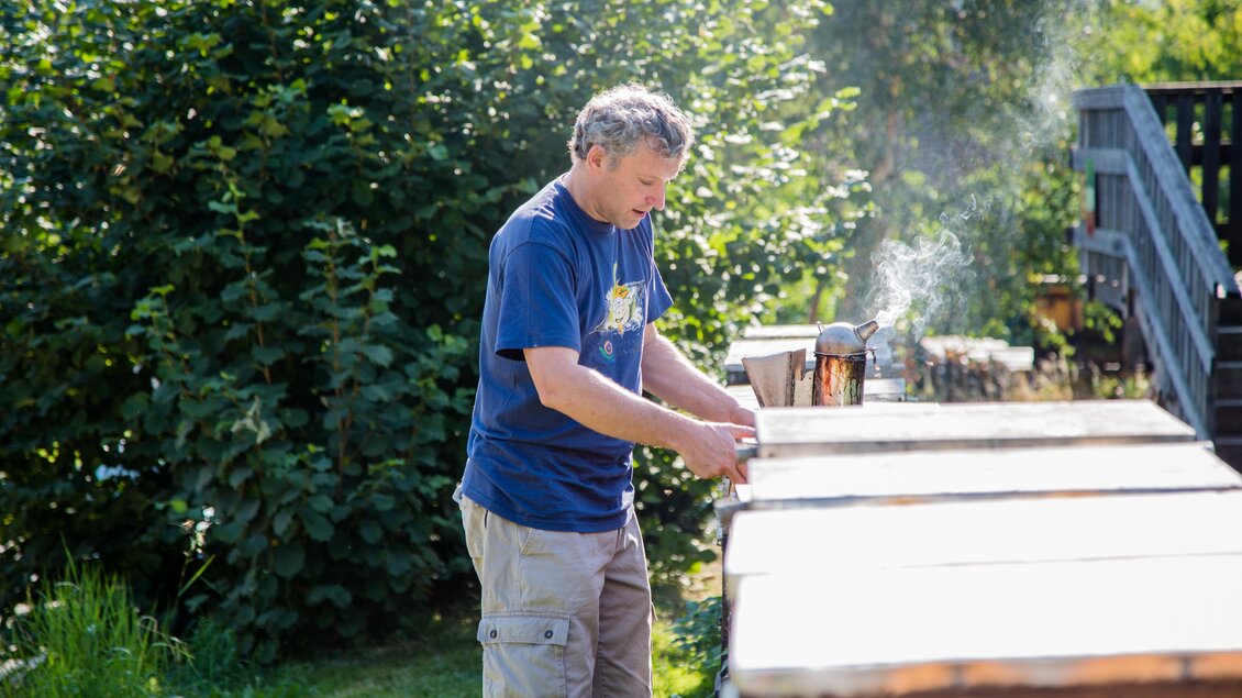 Ein Mann arbeitet im Garten an einem Holztisch. Im Hintergrund sind hohe Pflanzen und eine Treppe zu sehen.