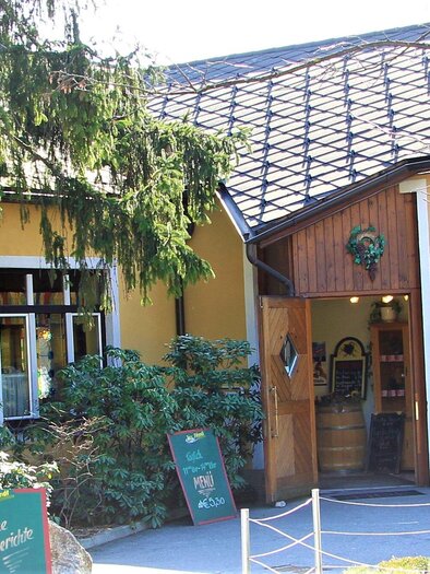 A cozy restaurant with a yellow facade and an inviting entrance. Surrounded by trees and green plants. | © In's Pfandl