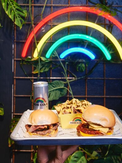 Serving tray with two burgers, fries with sauce, and Indie Cola in front of a neon wall. | © Indie Burger - Samuel Kuro