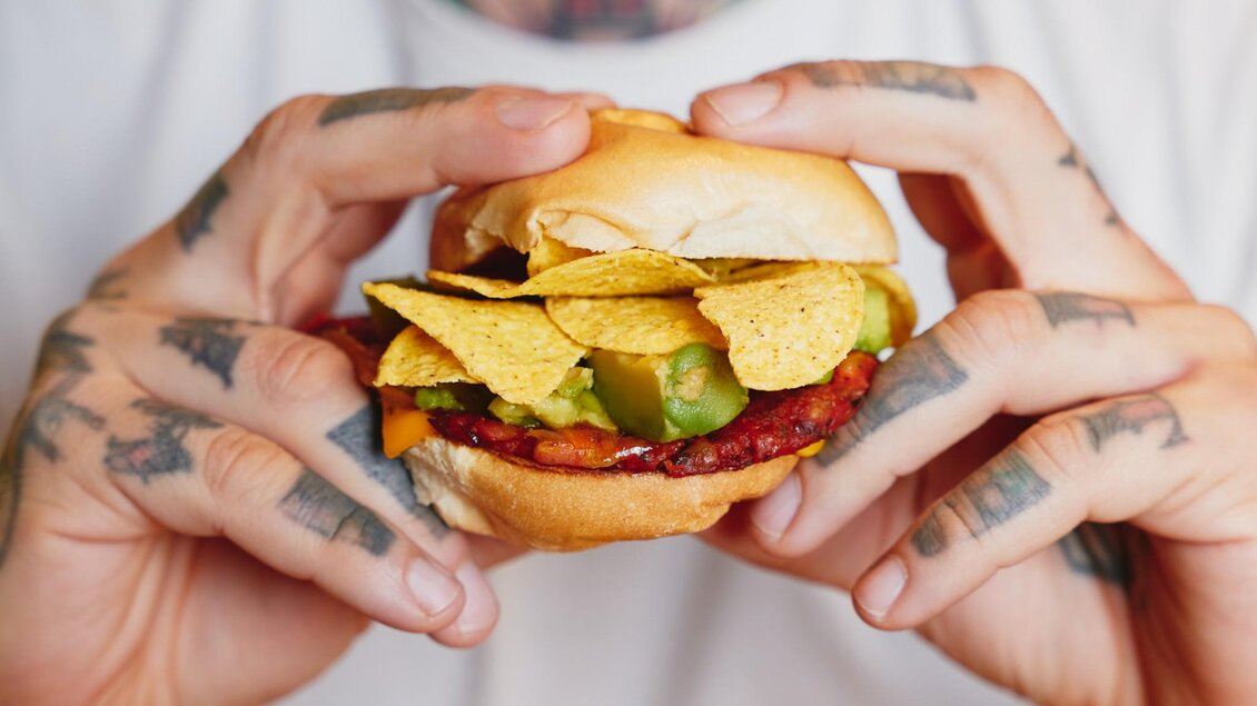 Nahaufnahme eines Burgers mit Nachos, Guacamole und Gemüse in tätowierten Händen. | © Indie Burger - Samuel Kuro