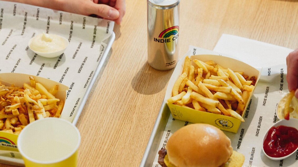Burger mit Nachos, Pommes, Ketchup und Indie Cola auf Tablett am Holztisch. | © Indie Burger - Samuel Kuro