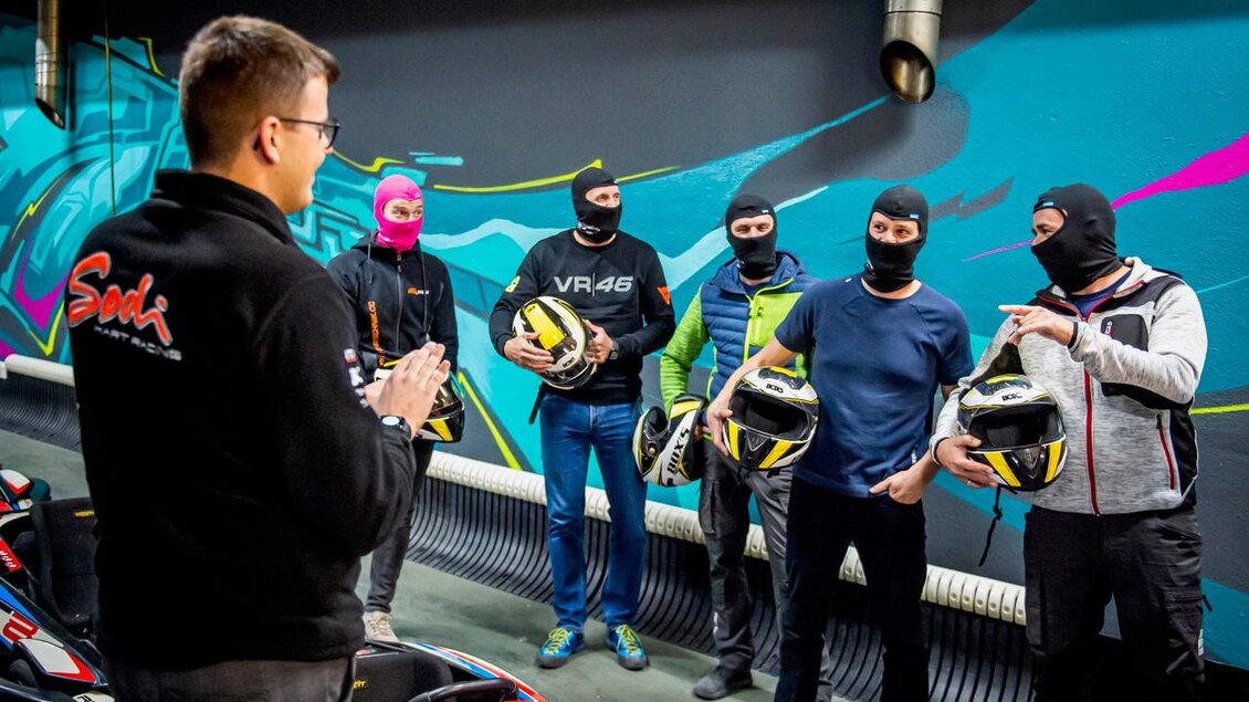 Eine Gruppe von Menschen in Schutzkleidung hört einem Sprecher zu. Sie halten Helme in der Hand und stehen vor Rennkarts in einer modernen Indoor-Rennstrecke. | © Indoorkart Spielberg