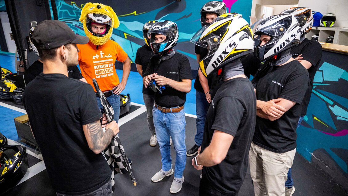 Eine Gruppe von Personen bespricht sich in einem Karting-Bereich. Einige tragen Helme, während eine Person mit einem auffälligen Kostüm im Hintergrund steht. | © Indoorkart Spielberg