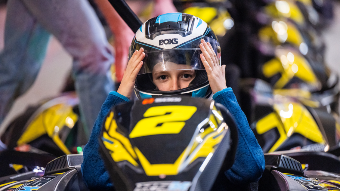 Ein Junge zieht seinen Helm an und sitzt in einem Go-Kart. Im Hintergrund sind mehrere Go-Karts in leuchtenden Farben aufgereiht. | © Indoorkart Spielberg