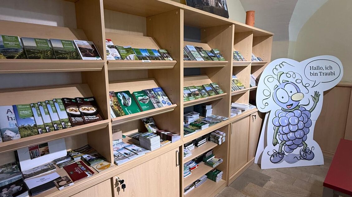 Ein gut organisierter Informationsbereich mit Regalen voll von Broschüren und Flyern. Eine lustige Weinfrau steht daneben und begrüßt die Besucher. | © TVB Südsteiermark