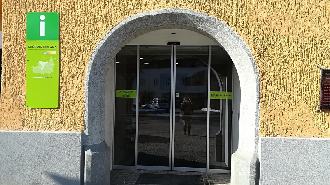 Ein Eingangsbereich mit einer Glas-Schiebetür und einer gewölbten Fassade. Links ist ein Informationsschild in grün mit einem Symbol. | © TVB Gröbminger Land 